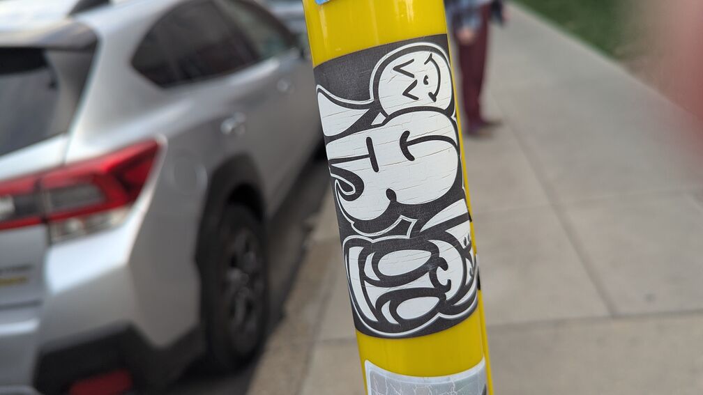 A graffiti slap on a pole featuring stylized bubble letters (that I can't make out) and the artist's signature big eye cartoon guy click for full resolution.