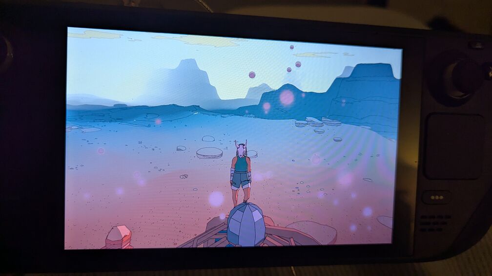 A screenshot from the videogame Sable. The main character stands on the top of a cliff, facing the camera. An otherworldly desert landscape stretches out behind her, in warm hues of red and blue. click for full resolution.