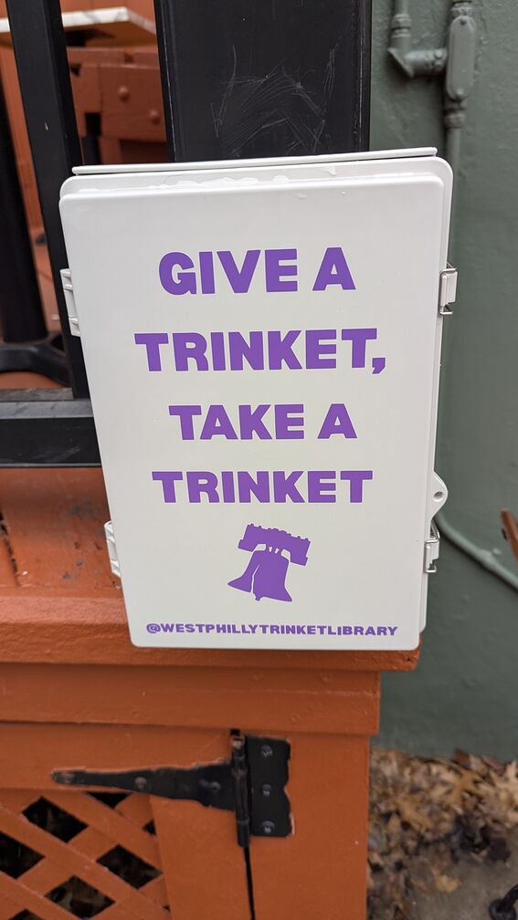 The West Philadelphia Trinket Library box, affixed to a post outside a coffee shop. The words 'Give a Trinket, Take a Trinket' are printed on it. click for full resolution.
