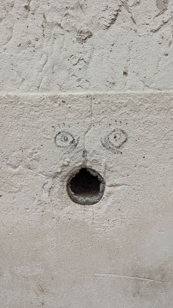 A round water drain in a wall in Lisbon, with some eyes drawn above it so it looks like a gaping face. click for full resolution.