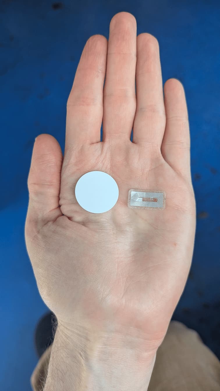 A photo of my hand, holding two different NFC tags. The first is a little white plastic disc around the size of a nickel, which has an NFC chip embedded in it. The second is a transparent sticker that looks like a circuit board -- this is a more clear example of what an NFC tag actually looks like. You can see the antenna that the chip uses to communicate and the teensy tiny computer that listens for messages.