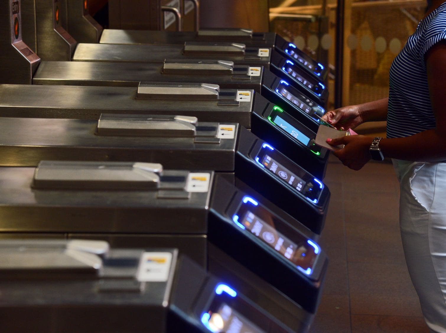 A row of New York City subway turnstiles, each with a glowing rectangle that you tap a credit card or phone against to unlock the gate. In the photo, a person is opening their wallet to pull out their card to press against the NFC reader.