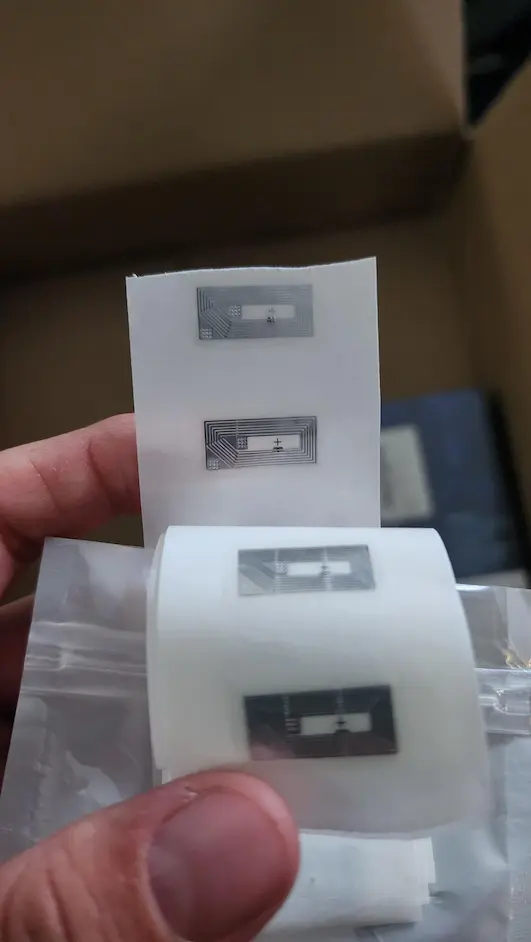 Somebody holding up a sheet of small NFC stickers. Each sticker is a fingernail sized silver rectangle.