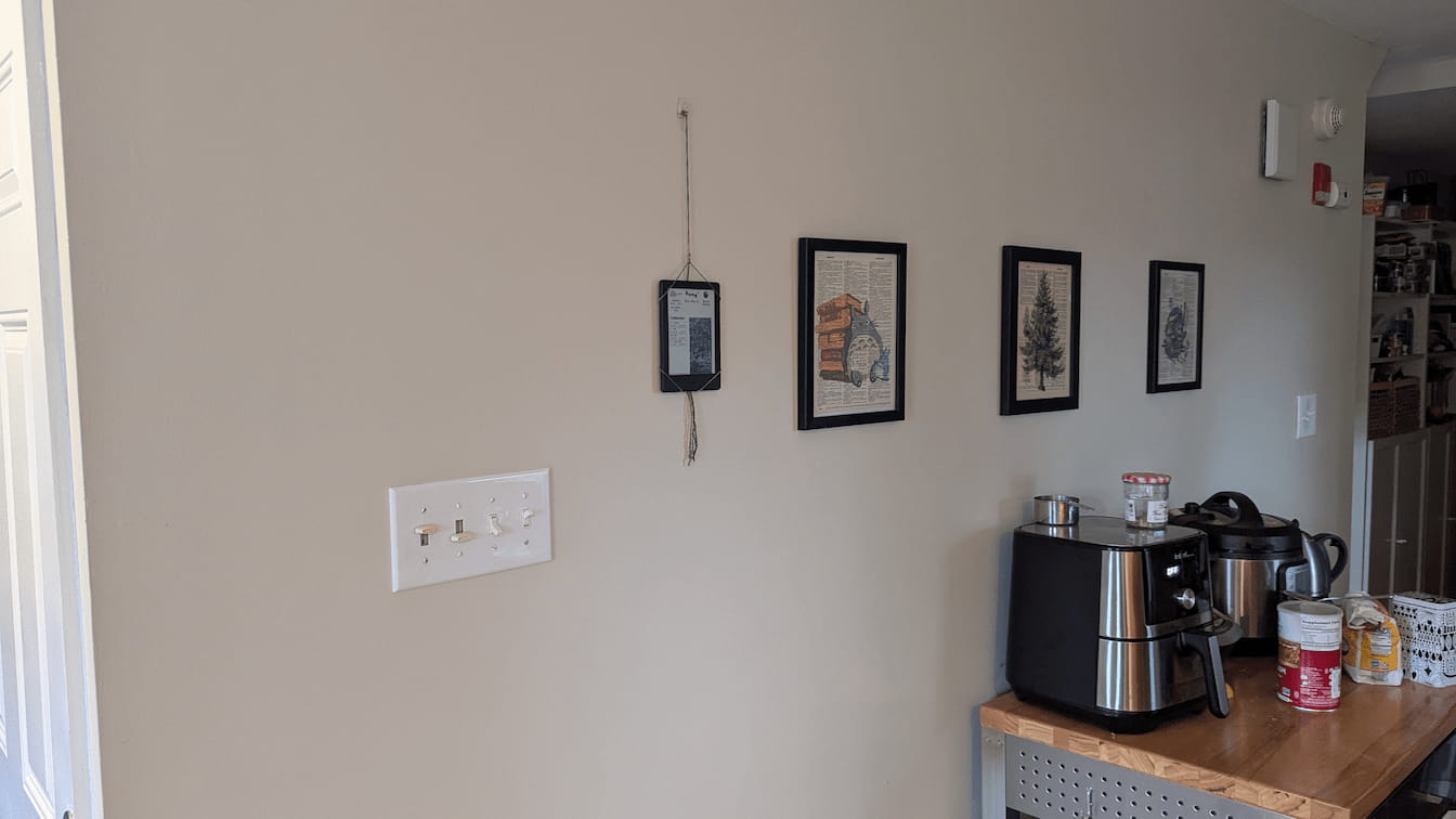 A photograph of my kitchen wall. The Kindle Display hang from a piece of string on end of the wall, followed by a series of framed Studio Ghibli illustrations. A kitchen island workspace sits against the wall, with an airfryer and instant pot on display.
