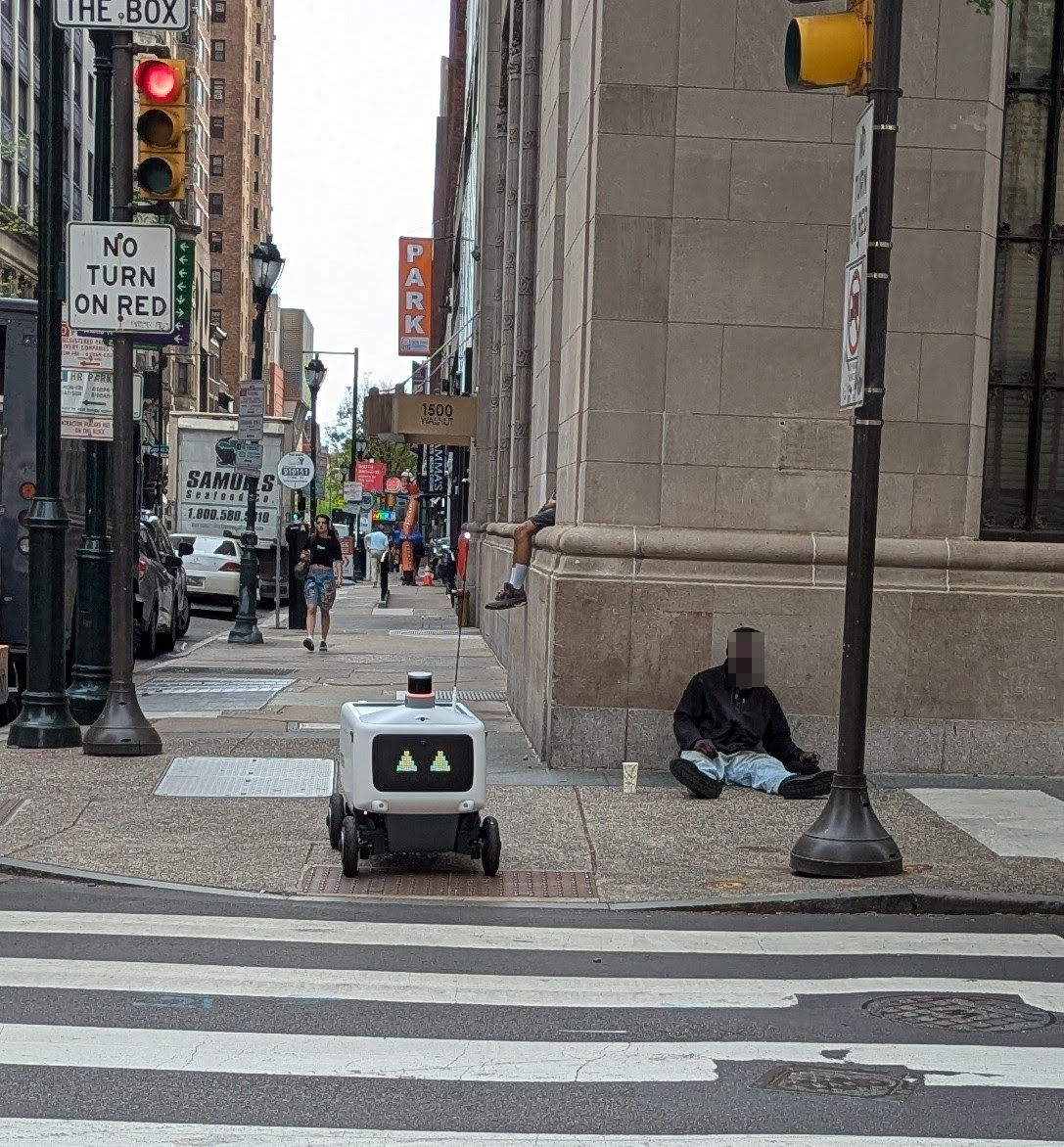 A photo of a busy street corner in Philadelphia.  An unhoused man is slumped against the wall of a building, with a paper cup next to him so he can panhandle. Meanwhile, 3 feet to his left is a food delivery robot, with smiling animatronic eyes, waiting to cross the street.