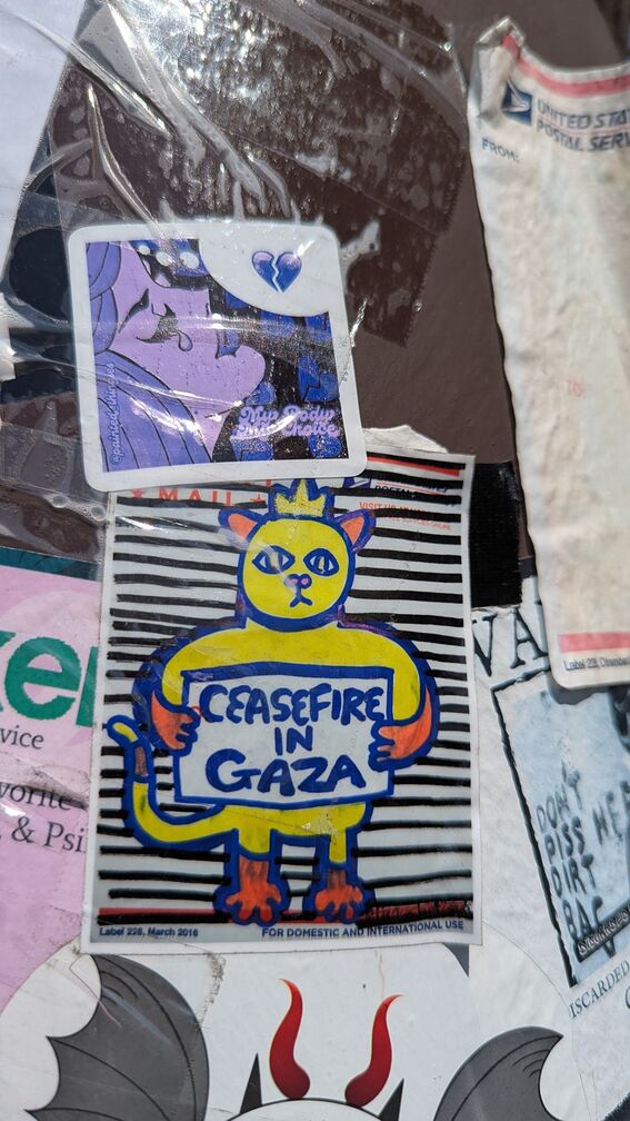 A slap drawn in marker on a USPS sticker. A yellow cat with a crown holds up a 'Ceasefire in Gaza' sign. A second sticker is above it, featuring a heartbroken cartoon woman and the words 'My Body My Choice'. click for full resolution.
