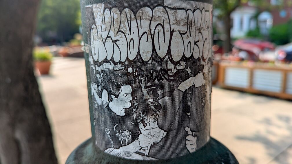 A dithered black and white photo of a fight at a punk show? Or a rally? One man is getting punched by another guy wearing a metal band shirt. There's a grafitti tag printed over the photo. Somebody has written the word hardcore in sharpie on the sticker. click for full resolution.