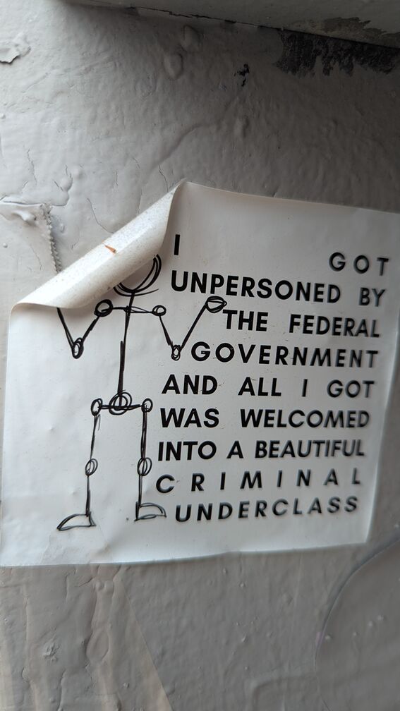 A sticker with a shrugging stick figure and text that reads: I got unpersoned by the federal government and all I got was welcomed into a beautiful criminal underclass. click for full resolution.