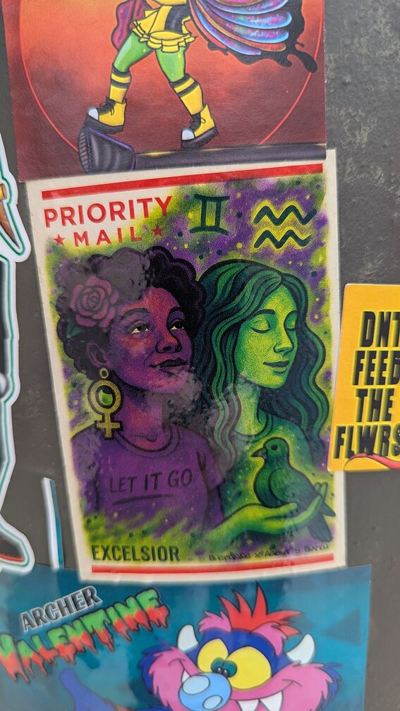 A priority mail stiker painted over with a drawing of two women. The first woman is black, with a giant female-sex sign earing, a red flower in her hair, and a tshirt thhat says: 'let it go'. She holds a pidgeon in her hand. The woman next to her is white, with her eyes closed, and long brown hair. I'm not quite sure what this all signifies, but it's beautiful. click for full resolution.